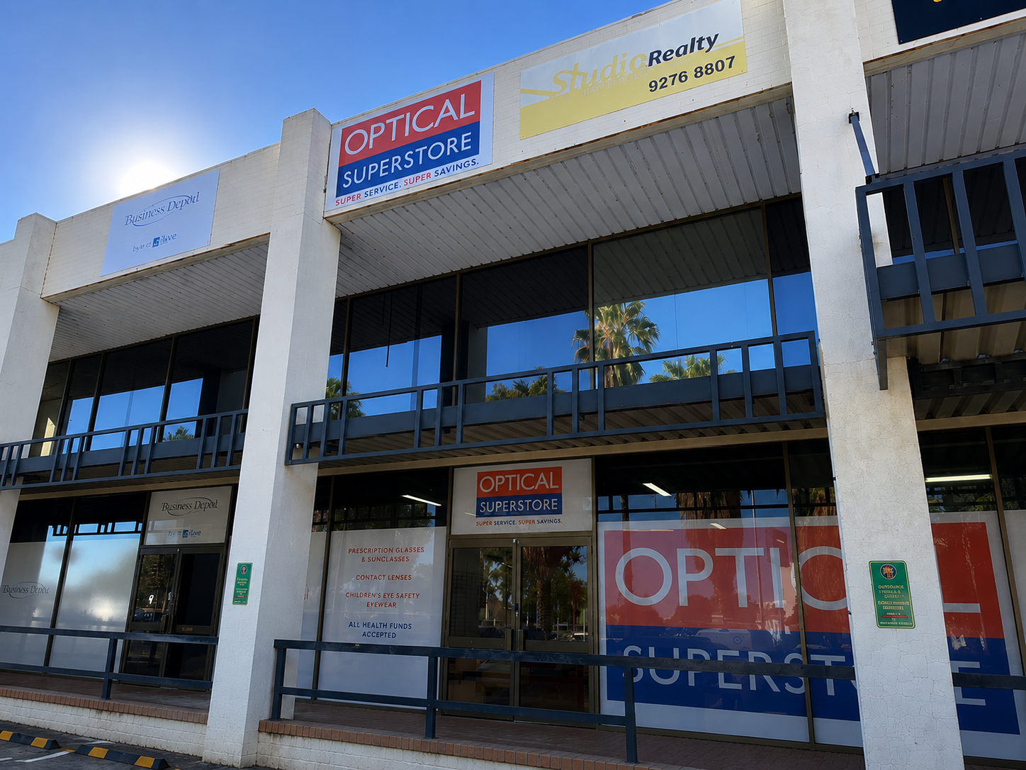 Facade of a strip mall optical shop with a large 'OPTICAL SUPERSTORE' sign above the entrance; glass storefronts and reflections of palm trees in the windows.