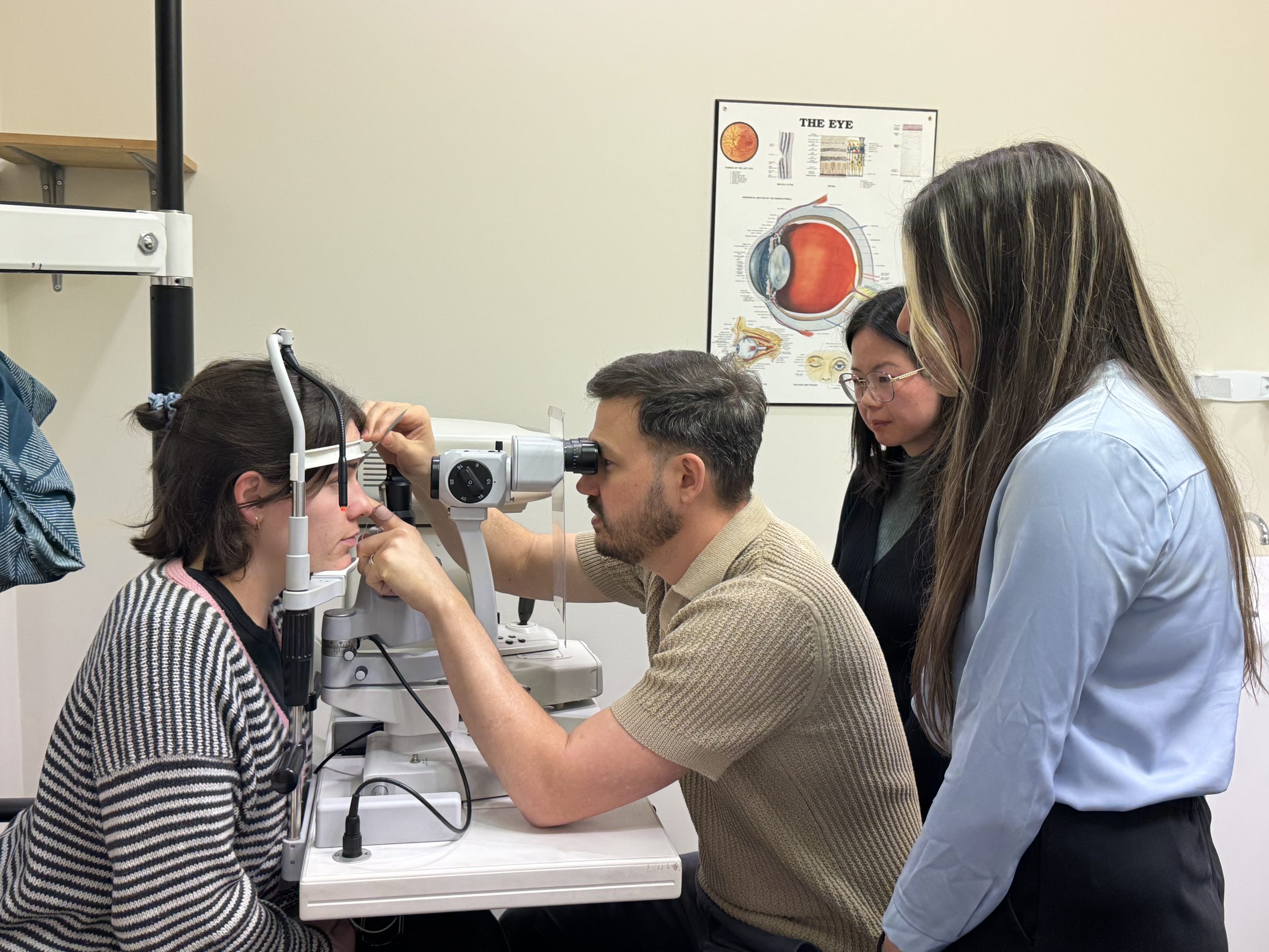Patient seated at a slit lamp undergoing an eye exam, clinician adjusting the instrument as another clinician stands nearby in a clinic with an eye poster on the wall