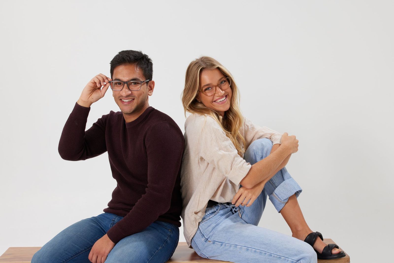 Two friends sit back-to-back on a wooden bench, smiling at the camera, wearing casual jeans and glasses.