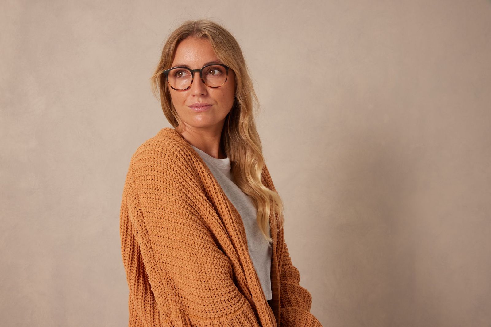 Woman with blonde hair and round glasses wearing an orange chunky cardigan, looking to her left against a beige backdrop.