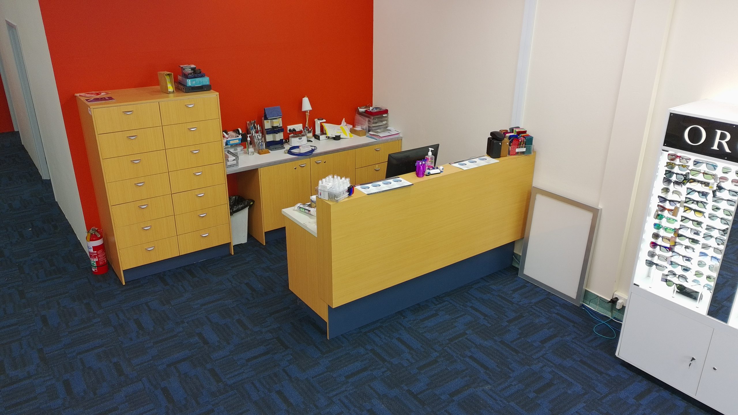 default - The Optical Superstore Optical shop counter area with wooden drawers, tools, and sunglasses on display near a bright orange wall and white cabinets.
