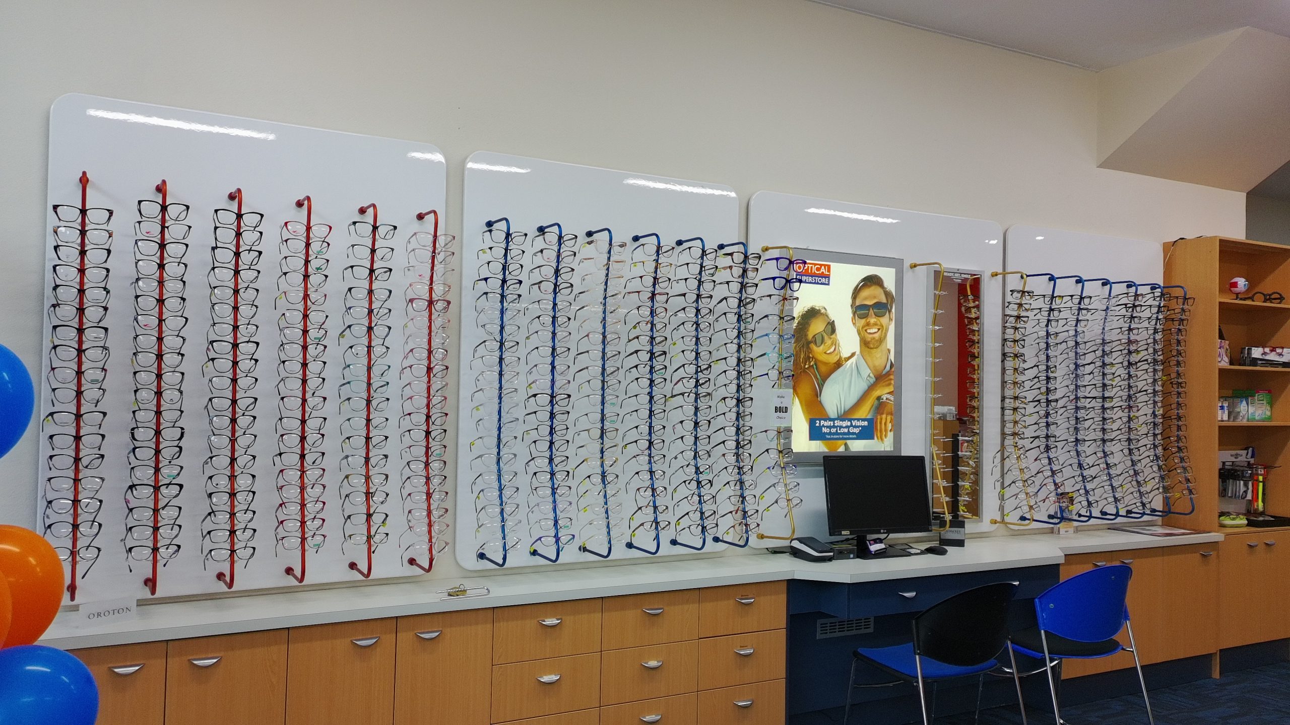 default - The Optical Superstore Eyewear store with wall-mounted displays holding rows of glasses in red, blue, and black frames next to a poster and shelves.