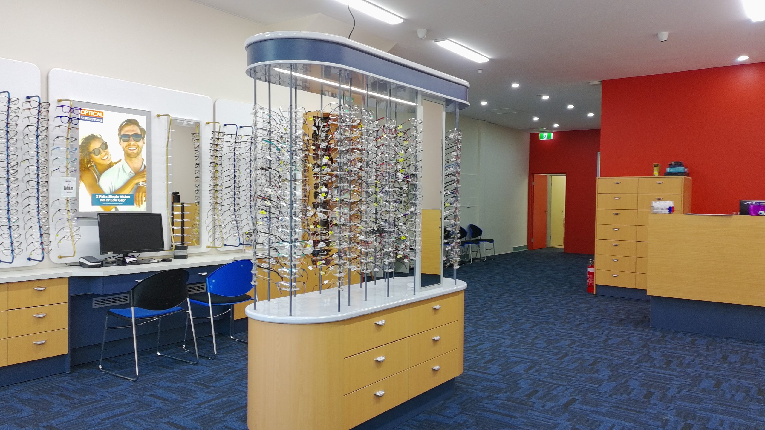 default - The Optical Superstore Interior of an optical store with rows of eyeglass display racks, a curved lighted central showcase, and a reception desk area against a red wall.