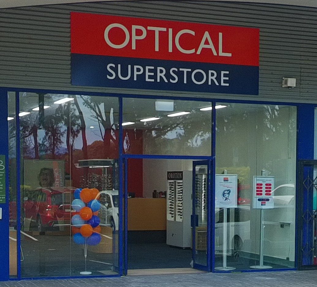 Optical Superstore storefront with red and blue sign and glass doors; blue frame and balloons inside.