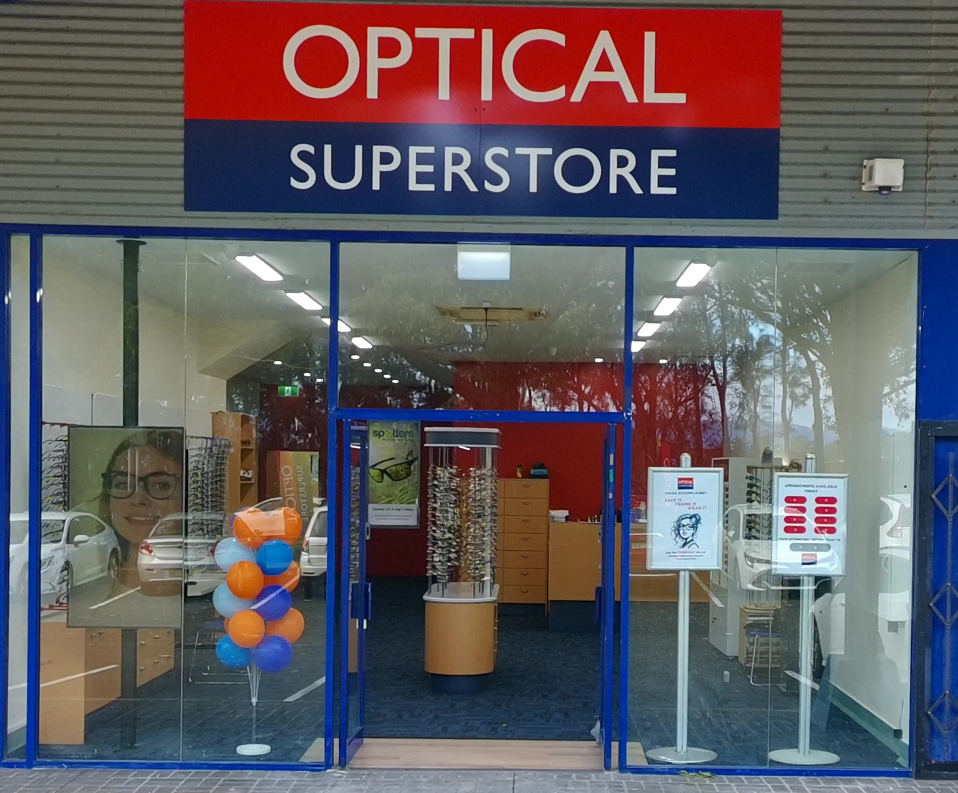Storefront exterior of an optical shop with a red-and-blue sign reading 'OPTICAL SUPERSTORE', blue-framed glass doors, and eyewear displays inside.