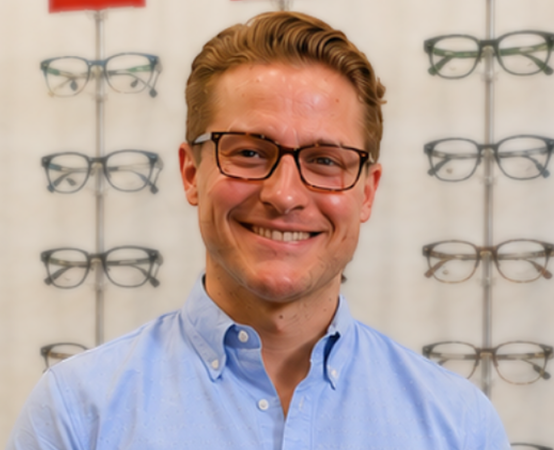 Smiling man wearing glasses in an eyewear store with display racks of glasses behind him from left to right.