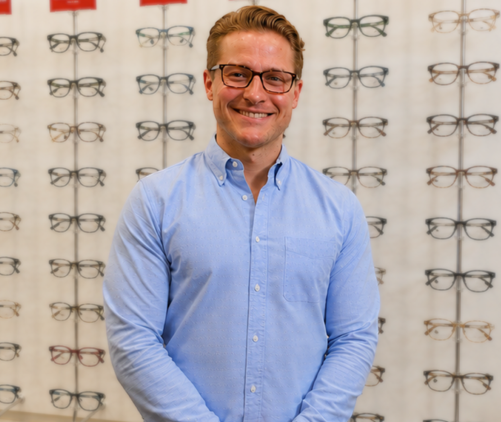 Smiling man in a blue button-down shirt stands in front of a wall of eyeglasses in an optical store while smiling for the camera.