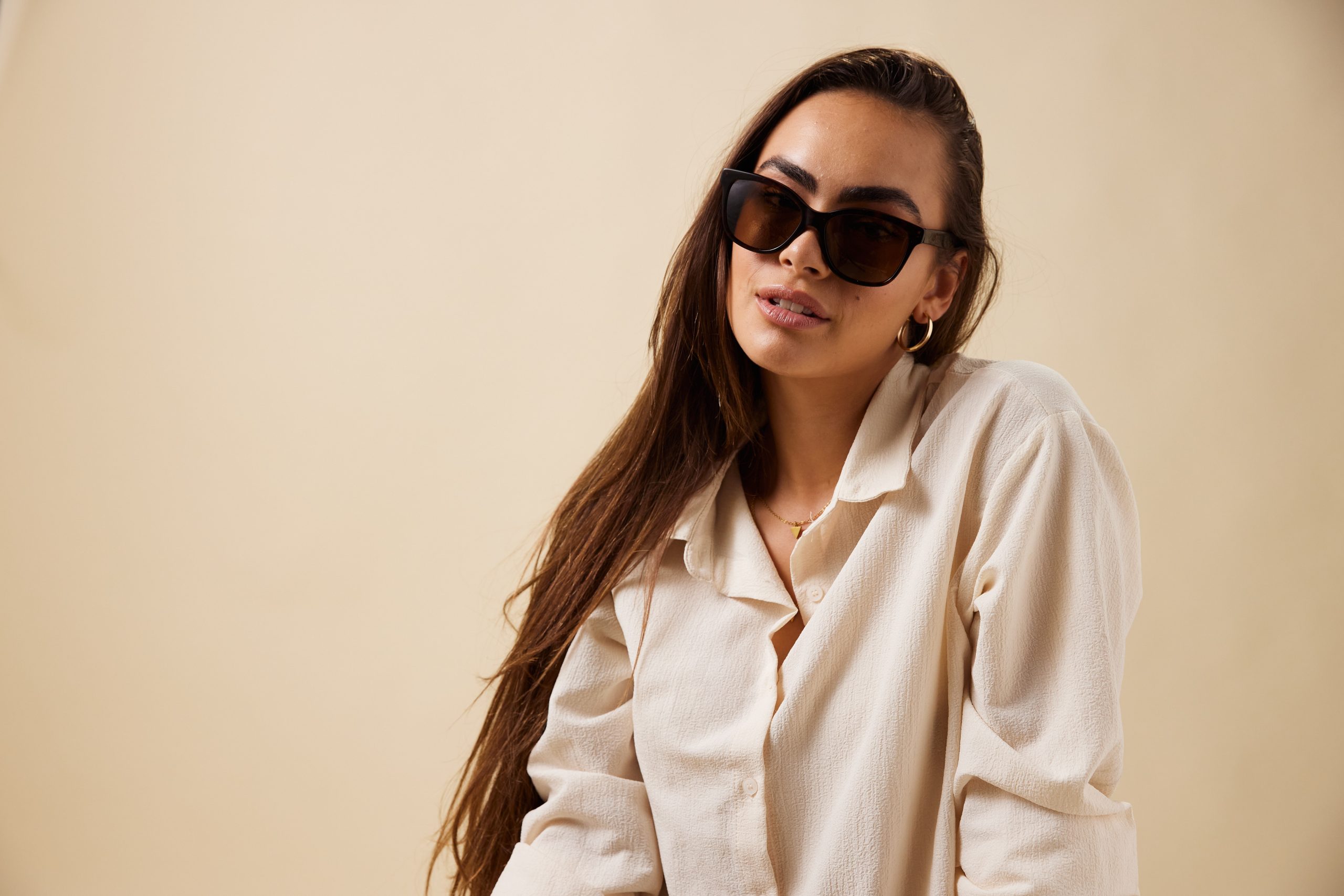 Woman wearing oversized black sunglasses and a beige button-up shirt, posing against a pale beige background.