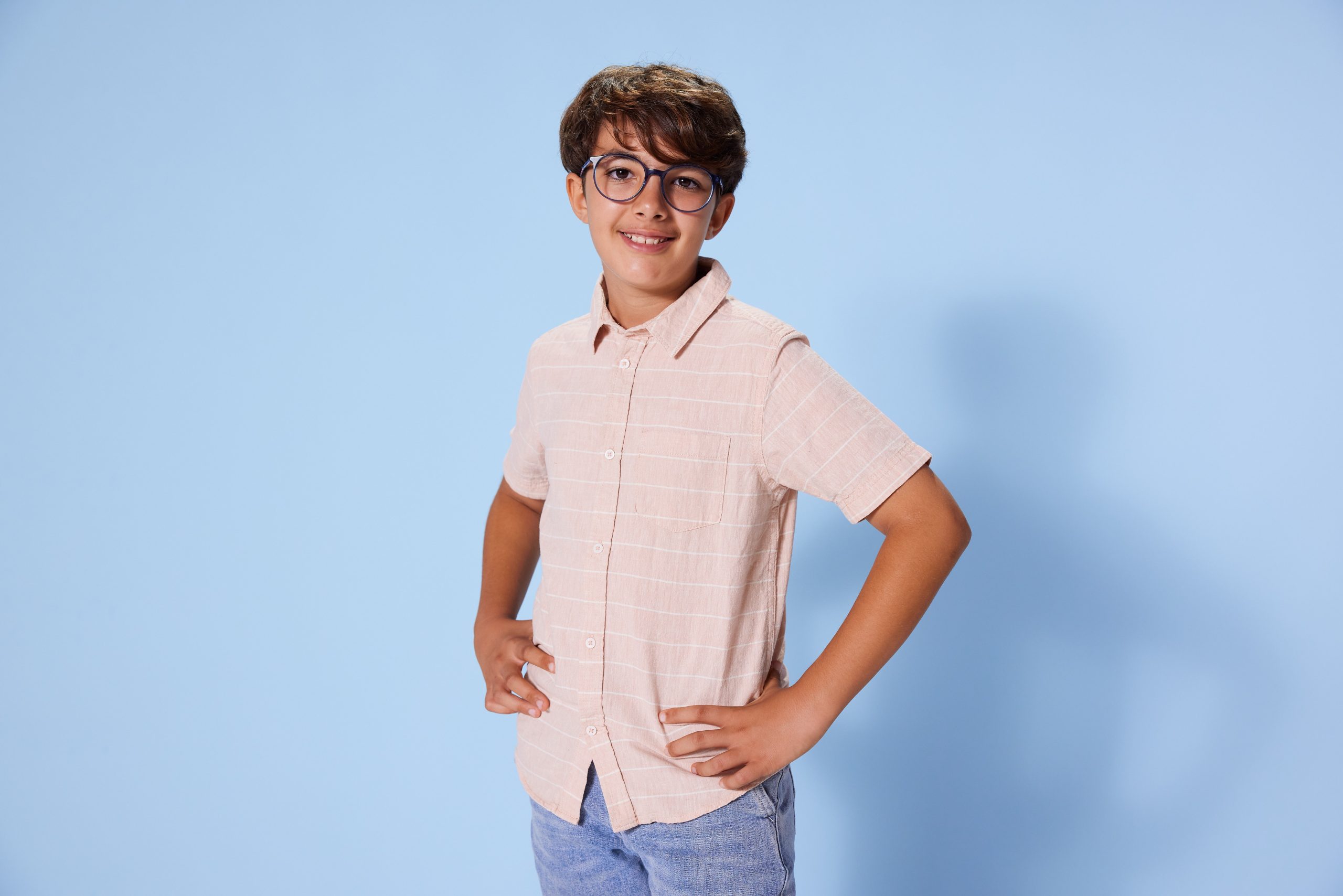 Smiling boy in a pale pink striped shirt and blue jeans stands with hands on hips against a light blue background.