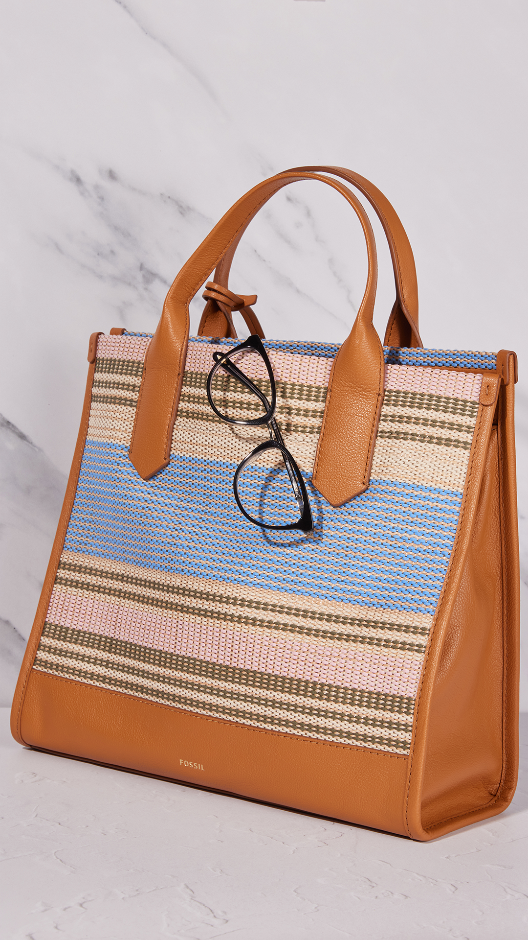 Tan leather-trimmed woven tote bag with blue and pastel stripes; black glasses hang from the handle.