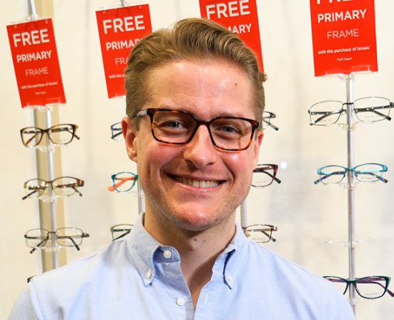 Smiling man wearing rectangular glasses in an eyewear shop with red sale signs behind him, blue button-down shirt visible.