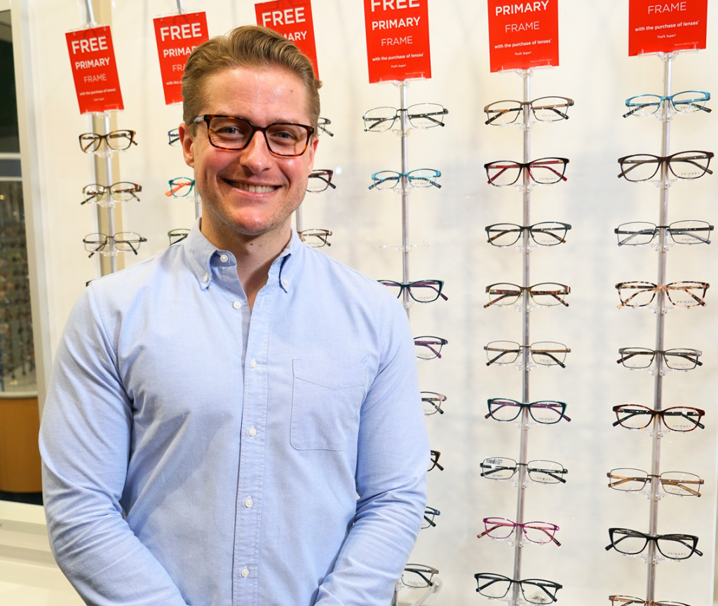 Smiling man in a light blue button-down shirt wearing glasses, standing in front of a wall of eyeglasses on display in an optical shop