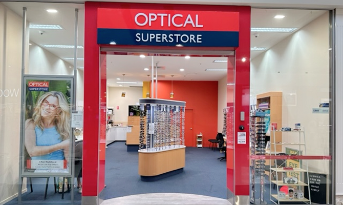 Entrance to an optical store with bold red frame and a blue 'SUPERSTORE' sign; eyeglasses displays visible inside the well-lit shop front.