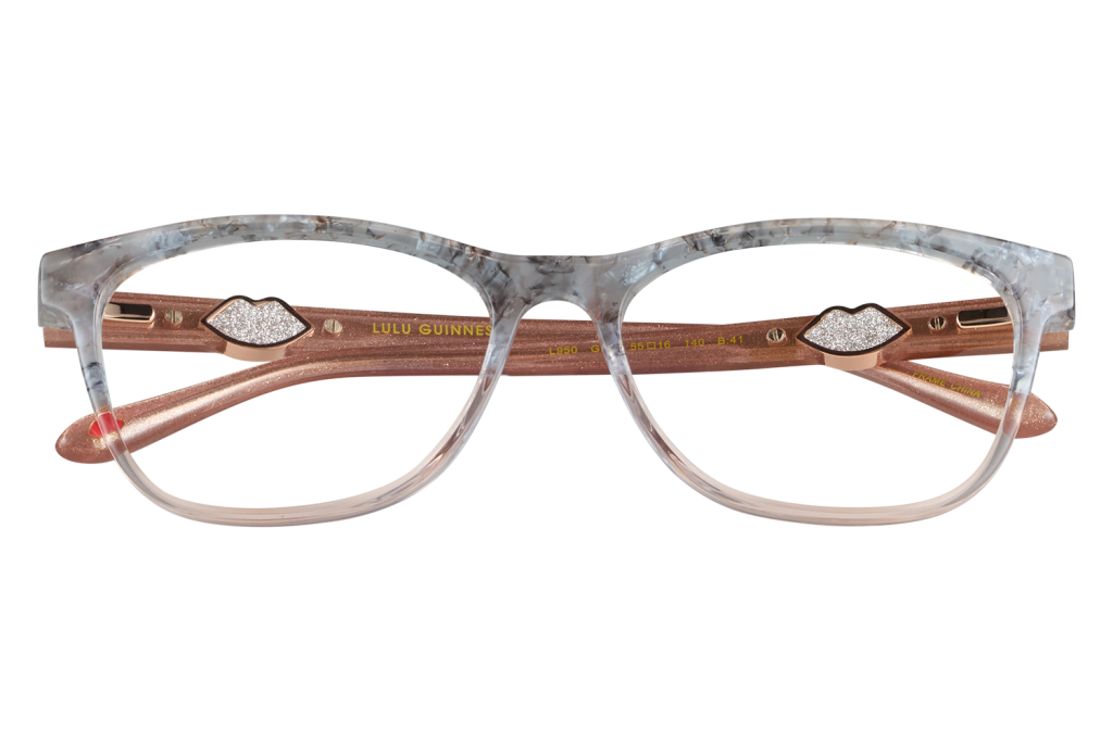 Pink marbled-frame eyeglasses with bronze temples and lip-shaped decorative accents resting on glass blocks, displayed on a gray background.