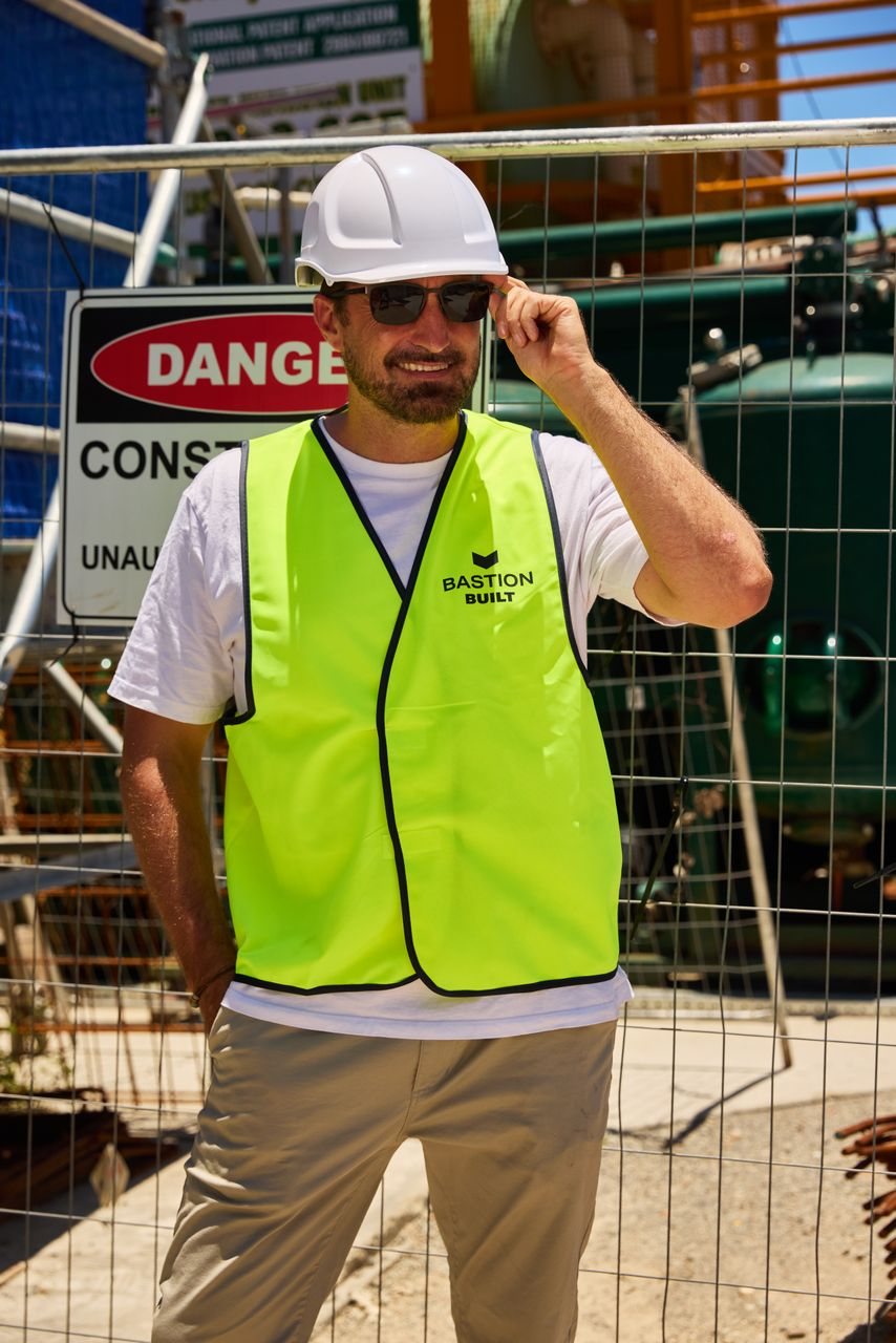 Construction worker in a bright neon safety vest and white hard hat standing behind a fence at a construction site with a danger sign, adjusting sunglasses.