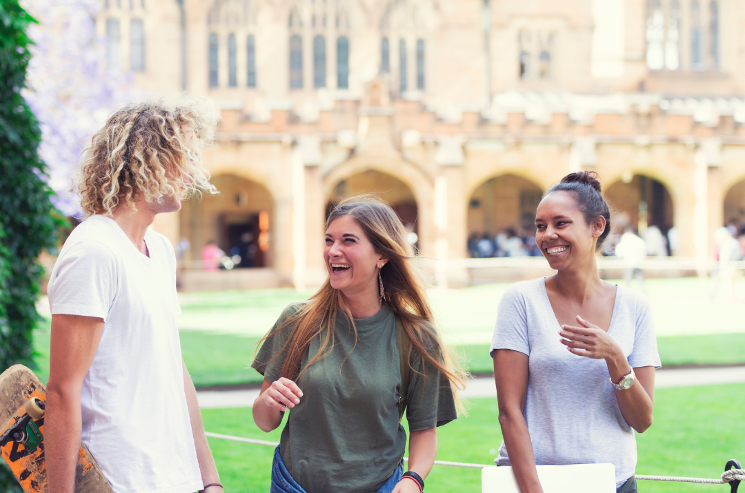 Three friends laugh and chat on a sunny college campus lawn, with one holding a skateboard nearby.