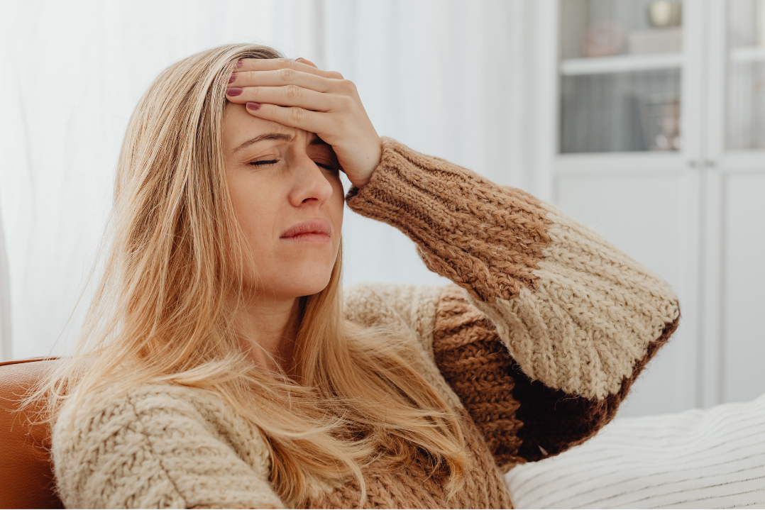 Woman sitting indoors, eyes closed, pressing her forehead with her hand to indicate a headache or fatigue.
