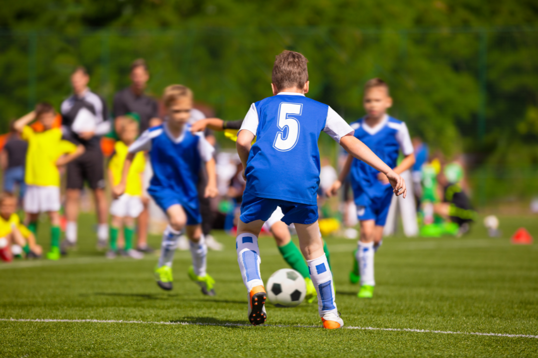 Youth soccer game: a player in blue jersey number 5 controls the ball on a green field as teammates close in nearby.